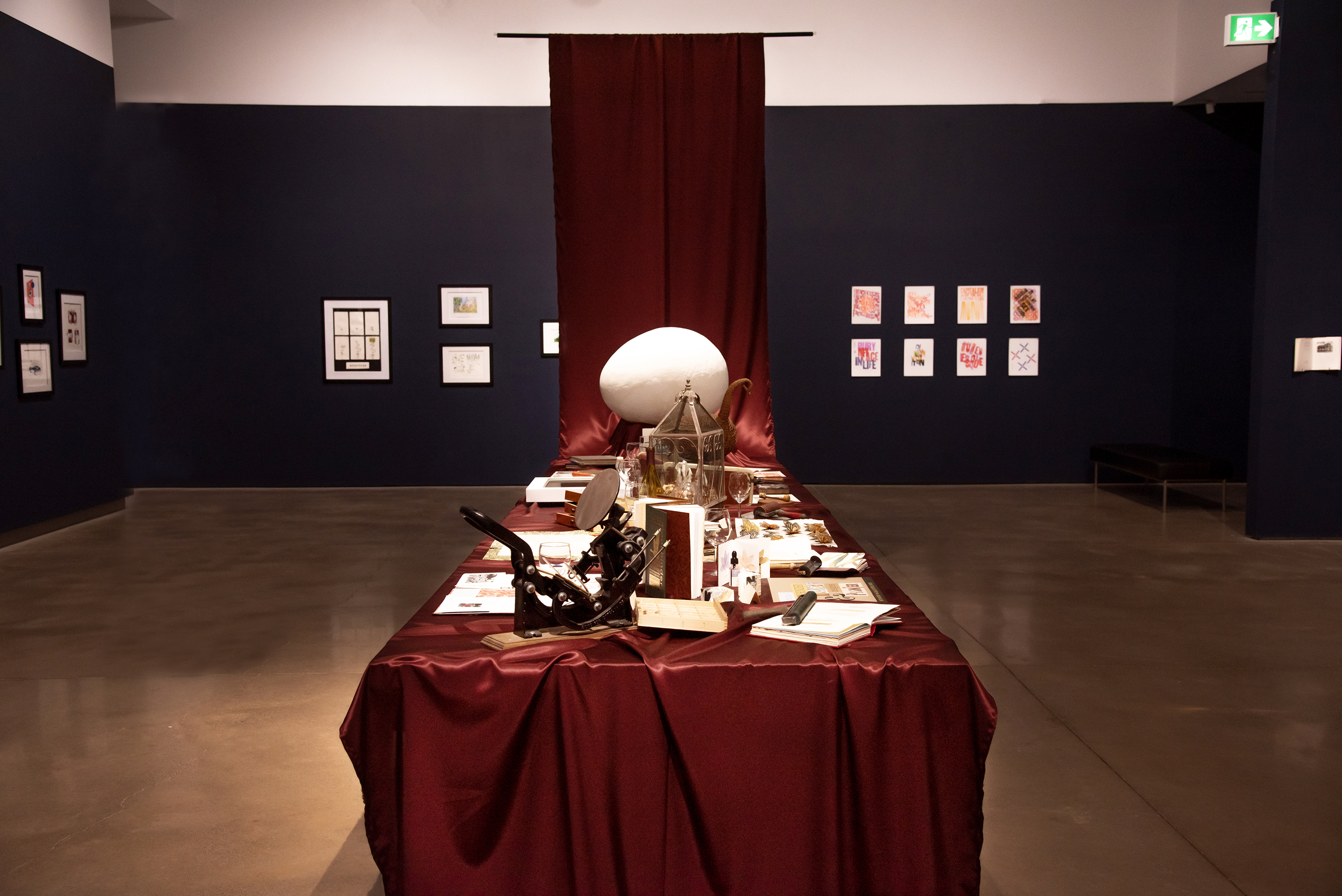 A banquet table draped in crimson satin which rises to the ceiling at one end.  Atop the table are various printmaking and book art work and ephemera