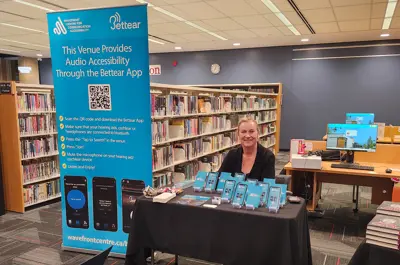 A woman smiling sat down at "Bettear" table, where people can get accessible listening accommodations for the event.