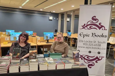 Two women sat behind a table of books. Their table sign reads "Epics Books" and they are vendors selling copies of books at our author series.
